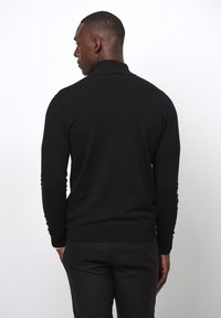 Schwarzer Strickpullover mit einem hohen Kragen, langen Ärmeln und gerippten Bündchen. Hat ein tailliertes Design und eine glatte Textur und wird über schwarzen Hosen getragen.