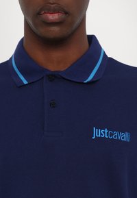 Mornarska polo majica z teksturirano tkanino, ki ima ovratnik z svetlo modrimi črtami in modrim vezenim logotipom na prsih.