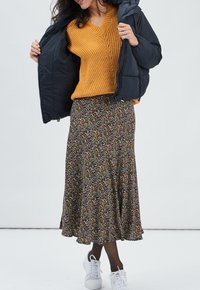 Gelbes Strickpullover mit V-Ausschnitt, schwarze Pufferjacke, Rock mit Blumenmuster und weiße Sneaker, die Textur und Schichten-Design hervorheben.