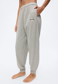 Άτομο που φοράει φαρδιά ανοιχτόγρια φόρμα με ελαστικά μανσέτες και λογότυπο Calvin Klein στο πάνω μέρος του μηρού, όρθιο ξυπόλητο σε απλό φόντο.