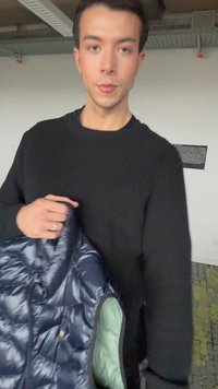 Νεαρό άτομο με μαύρο πουλόβερ που κρατάει ένα σκούρο μπλε μπουφάν μέσα σε κλειστό χώρο, στέκεται μπροστά σε λευκό τοίχο με πινακίδα κειμένου.