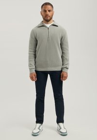 Lichtgrijze geribde trui met een halve rits kraag, gecombineerd met donkere jeans en witte sneakers. Textuur van de stof zichtbaar op de trui.