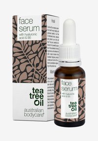 Brun glassflaske med hvit pipette, merket "ansiktsserum" med tea tree olje, hyaluronsyre og B5. Boks med abstrakt brunt og kremmønster.