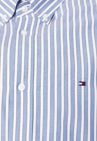 Blauw-wit gestreept overhemd met knopen en ronde kraag, met witte knopen en een klein geborduurd logo in rood, wit en blauw.