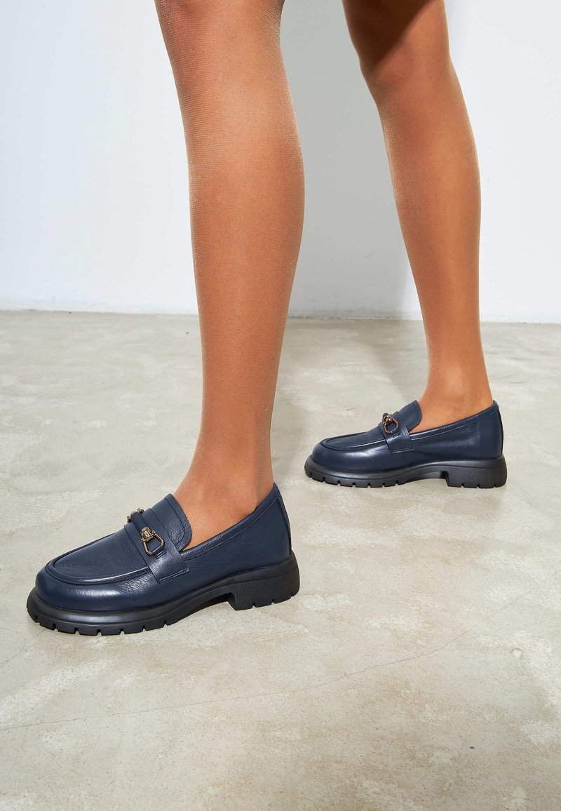Mörkblå läderloafer med texturerad yta, som har en metallackcent och en kraftig svart sula. Slät ovandel med rundad tådesign.