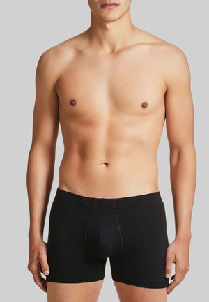 Torso maschile con boxer neri, braccia rilassate lungo i fianchi, su uno sfondo grigio uniforme.