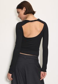 Rövid, sötét hajú nő fekete, nyitott hátú, hosszú ujjú felsőt és sötét csíkos nadrágot visel, egy sima fehér falnak támaszkodva.