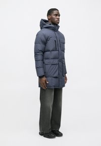 Ναυτικό χρώμα padded parka μπουφάν με κουκούλα, πολλαπλές τσέπες και λεία υφή. Φορεμένο με φαρδιά σκούρα τζιν και μαύρα παπούτσια.