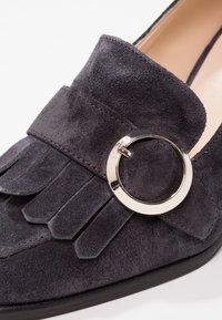 Mocassin en daim gris foncé avec une large bande ornée de franges décoratives et une boucle circulaire argentée brillante. Doublure intérieure lisse.