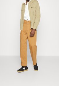 Chaqueta beige con botones y dos bolsillos en el pecho, camisa blanca debajo, pantalones naranja-marrón y zapatillas negras con detalles en blanco.