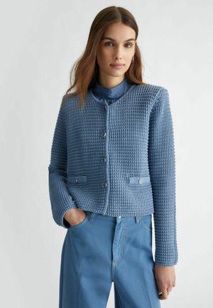 Giacca blu testurizzata con scollo rotondo, chiusura a bottoni e due tasche frontali, abbinata a pantaloni in denim blu chiaro.