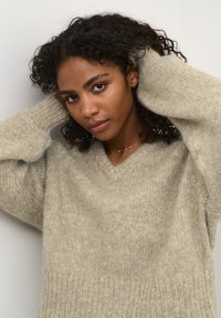 Pull en maille oversize beige clair avec un décolleté en V, un tissu texturé et des manches larges. Le mannequin pose les mains derrière la tête contre un fond neutre.