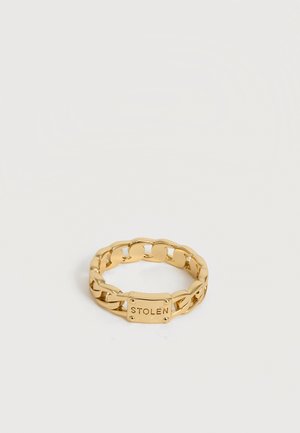 Χρυσό δαχτυλίδι με αλυσίδα και ορθογώνια πλακέτα με χαραγμένη τη λέξη "STOLEN" σε λευκό φόντο.