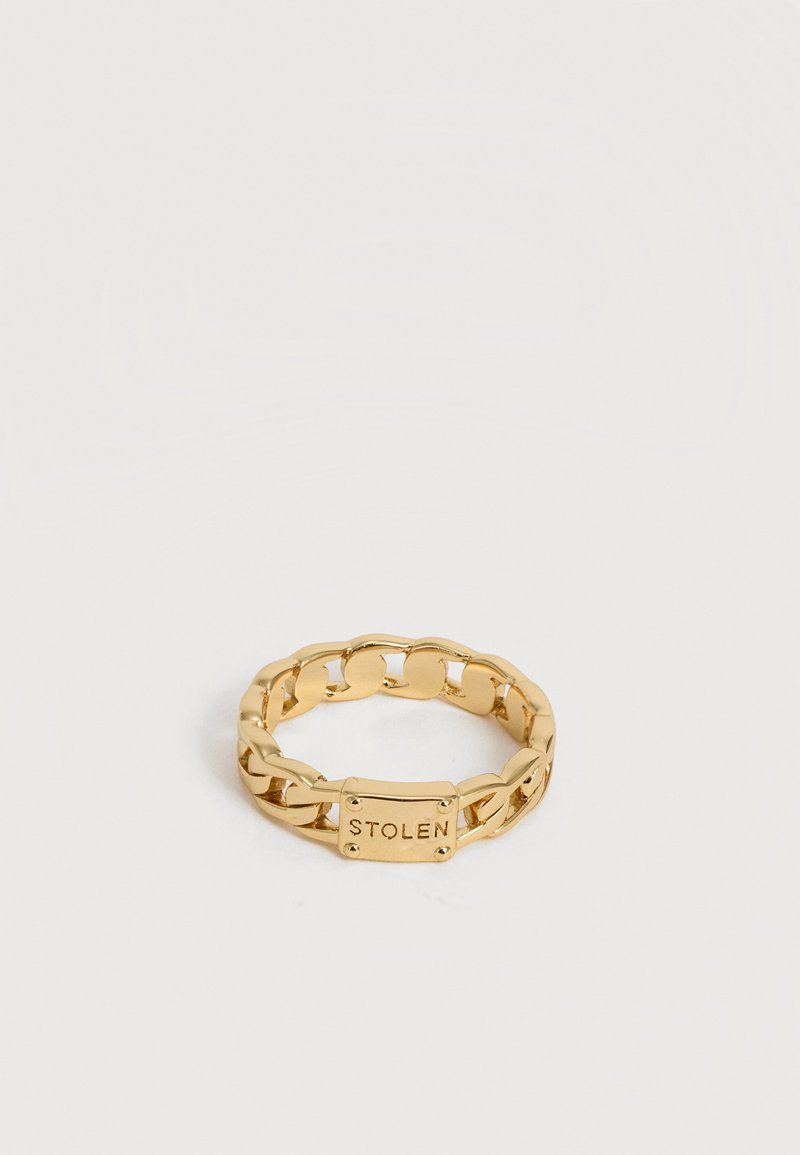 Χρυσό δαχτυλίδι με αλυσίδα και ορθογώνια πλακέτα με χαραγμένη τη λέξη "STOLEN" σε λευκό φόντο.