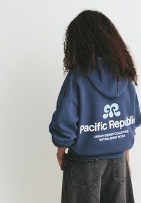 Námornícky modrá mikina z jemného materiálu s veľkým bielym grafickým prvkom a textom "Pacifická republika" a "URBAN DESIGN COLLECTION ZALOŽENÁ V ROKU 2018."