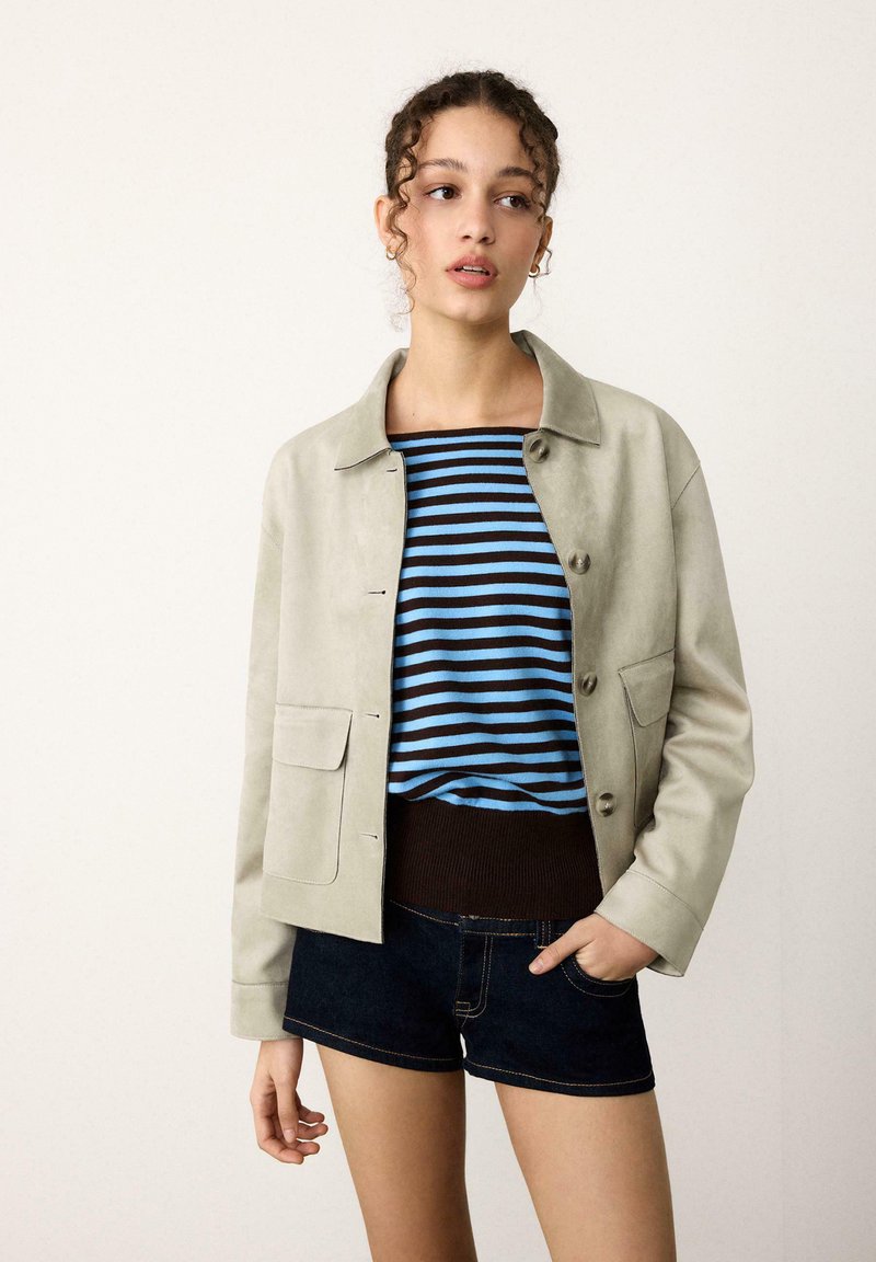 Veste en daim vert clair avec de grandes poches avant, associée à un pull rayé bleu et marron et à un short en denim foncé.