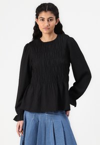 Black long-sleeve blouse with textured smocking detail, featuring a round neckline and gathered waist, paired with a denim pleated skirt.