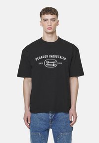 Fiatal férfi fekete pólóban, amin a "Pegador Industries Since 2018" felirat és egy kulcs grafika látható, kék farmernadrággal párosítva.