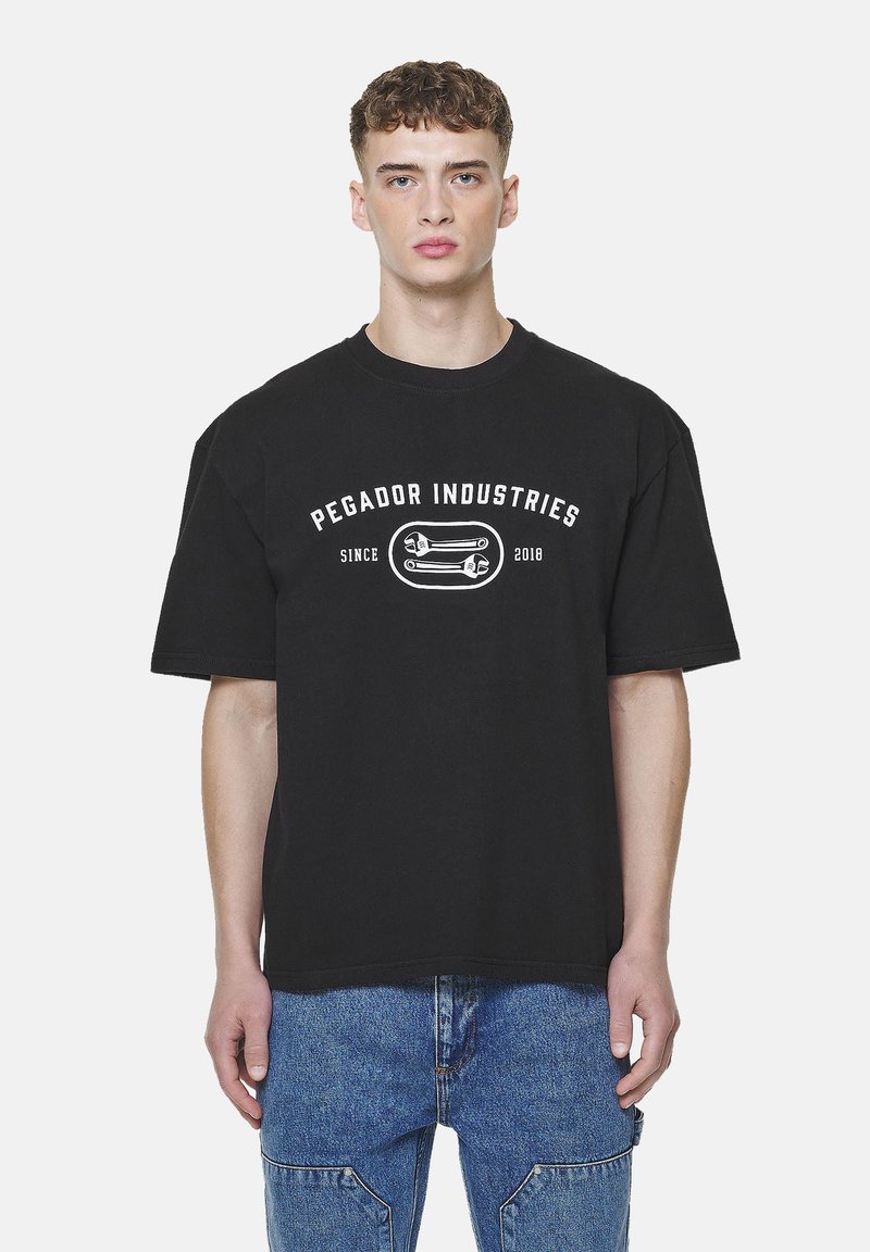 Fiatal férfi fekete pólóban, amin a "Pegador Industries Since 2018" felirat és egy kulcs grafika látható, kék farmernadrággal párosítva.