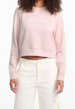Femme portant un sweat court rose clair avec le texte "Guess Jeans" et un pantalon cargo blanc taille haute, debout, les bras détendus le long du corps.