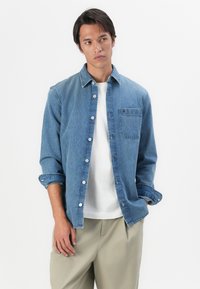 Lichtblauwe denimshirt met knoopsluiting aan de voorkant, borstzak en opgerolde mouwen. Zachte textuur, casual pasvorm en ronde kraag.