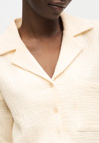 Κοντινό πλάνο ενός ατόμου που φοράει ένα κρεμ πουκάμισο με υφή, κουμπιά, γιακά και τσέπη στο στήθος.