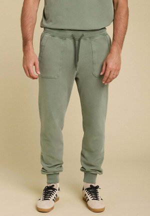 Joggers en coton verts avec poches latérales et chevilles resserrées, dotés d'une taille élastique avec cordon de serrage. Portés avec des baskets beiges et des lacets noirs.