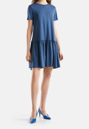 Kobieta ubrana w niebieską sukienkę z krótkimi rękawami sięgającą kolan, z marszczonym dołem oraz niebieskie szpilki na szpic, stojąca na białym tle.