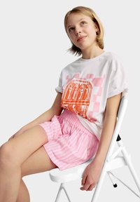 Witte T-shirt met oranje grafische print, gecombineerd met roze gestreepte shorts. Zit op een witte stoel, met kort, steil haar.