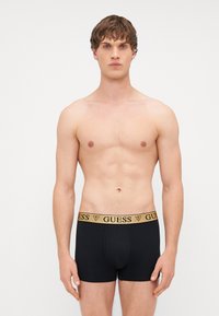 Melnu vīriešu bokseršortu priekšauts ar zelta "GUESS" logotipu uz jostas. Audums izskatās mīksts un elastīgs. Pārsēdēšana ir cieša un formveidīga.