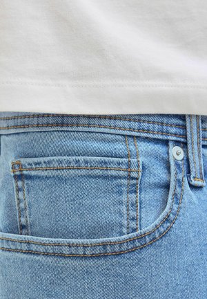 Približni prikaz stražnjeg džepa svijetloplavih teksas hlača s bijelom košuljom uvučenom iznad pojasa.