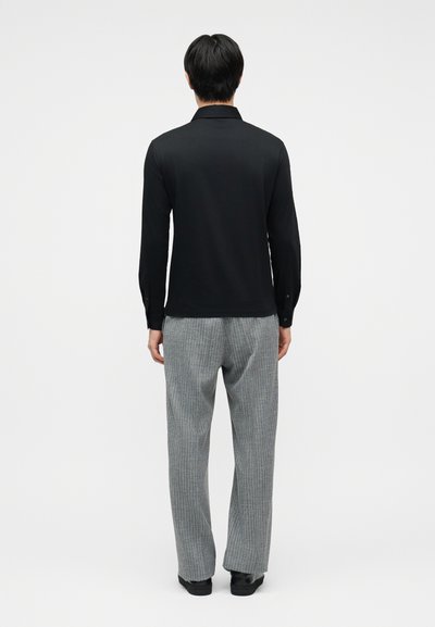 Άνδρας που φοράει μαύρο μπλουζάκι μακρυμάνικο και γκρι παντελόνι με ριγέ σχέδιο, στέκεται γυρισμένος προς τα πίσω με φόντο έναν απλό λευκό τοίχο.