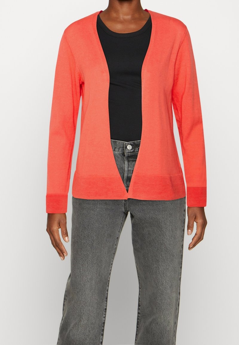 Koralfärgad kofta med V-ringning, ribbade ärmmuddar och öppen framsida. Bärs över en svart topp och matchas med grå jeans. Slät textur.