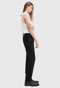 Pantalones de mezclilla negros con un diseño de pierna recta y estilo de cinco bolsillos. Llevados con una blusa sin mangas blanca y botas negras hasta el tobillo.