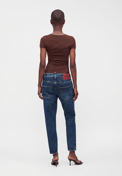 Personne aux cheveux courts porte un t-shirt marron ajusté, un jean bleu et des sandales marron à talons hauts, vue de dos sur un fond uni.
