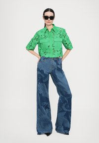 Green short-sleeve shirt with cut-out floral patterns and two front pockets, paired with wide-leg denim jeans with floral prints.