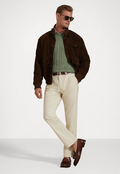 Καφέ σουέντ τζάκετ, πράσινη πλεχτή μπλούζα, μπεζ παντελόνι, καφέ δερμάτινα loafers. Περιλαμβάνει γυαλιά ηλίου; απλός σχεδιασμός χωρίς ορατά σχέδια.