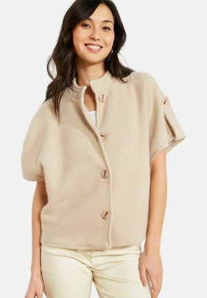 Frau trägt eine beige strukturierte, kurzärmelige, geknöpfte Strickjacke mit Stehkragen über einer hellen Hose, lächelt mit lockerem dunklem Haar.