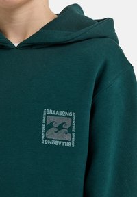Zelená mikina s kapucí vyrobená z měkké tkaniny, která na levé části hrudi nese čtvercové logo s nápisem "Billabong" a designem vlny v stříbrné barvě.