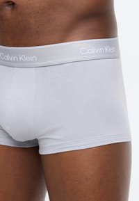 Κοντινό πλάνο ενός ατόμου που φοράει ανοιχτό γκρι μποξεράκια Calvin Klein, εμφανίζοντας τη ζώνη μέσης και το πάνω μέρος του μηρού.