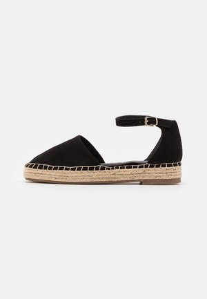 Sort espadrilles med flad sål, lukket tå, ankelrem med guldfarvet spænde og vævet jutesål på hvid baggrund.