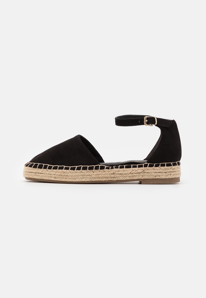 Chaussure plate espadrille noire avec bout fermé, bride de cheville avec boucle dorée et semelle en jute tissée sur fond blanc.