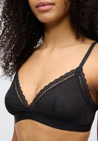 Mustast pitsist ääristatud bralette, mis on valmistatud läbipaistvast kangast õrnalt logo mustriga, V-kaelusega ja elastse logoga detailidega ribaga.