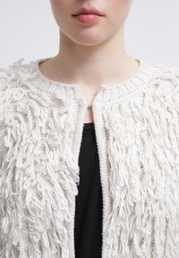 Vit texturerad cardigan med en loopad garndesign, rund halsringning och öppen framsidan. Bärs över ett svart plagg, vilket skapar kontrast.