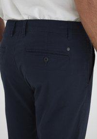 Navy blue pants made of cotton, featuring a back pocket with a button closure and a subtle logo detail near the waistline.