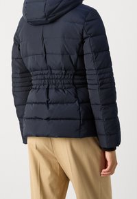 Veste matelassée bleu marine avec taille cintrée et design matelassé horizontal. Dotée d'une capuche et de poignets élastiqués pour un ajustement parfait.