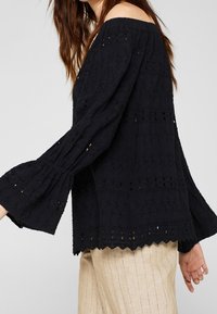 Sort off-shoulder top i struktureret stof, med klokkede ærmer og bølget kant, prydet med øjeletmønstre og detaljerede stikninger.