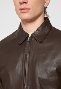 Giacca in pelle marrone con chiusura a zip, colletto a punta e texture liscia, caratterizzata da una leggera lucentezza e cuciture dettagliate.