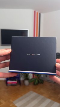 Scatola blu scuro Tommy Hilfiger con logo centrato sul davanti, con una striscia bianca e rossa sul bordo inferiore. Design rettangolare semplice.