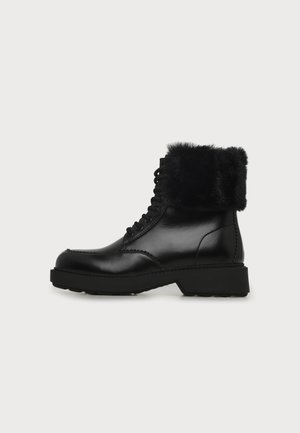 Bottine noire en cuir avec laçage à l'avant et revers en fausse fourrure, semelle épaisse et petit talon sur fond clair uni.